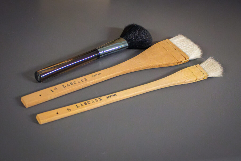 Närbild av tre penslar som används vid rengöring av verk. Den översta har ett skaft av plast och metall samt en rund, mörkfärgad borst. De två nedersta penslarna är platta och av trä, med platta vita borst.