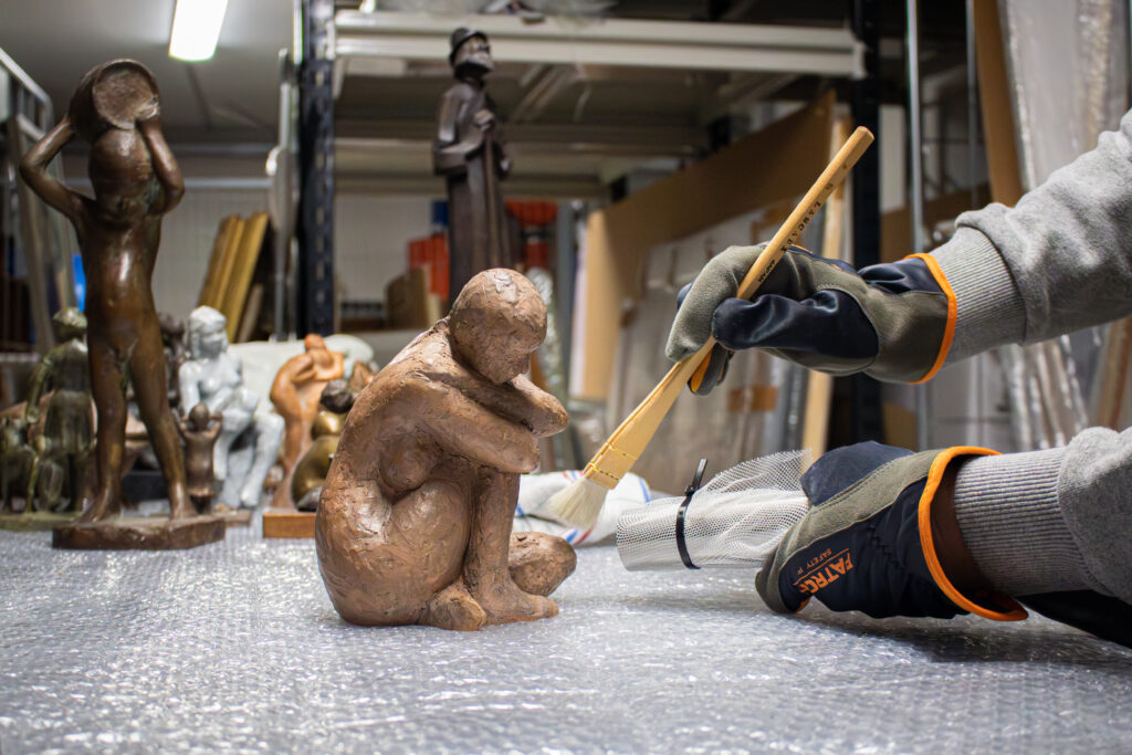 Närbild av rengöringen av en skulptur i röd lera med hjälp av pensel och dammsugare. Verket föreställer en hopkrupen naken kvinna och ligger på ett bord.
