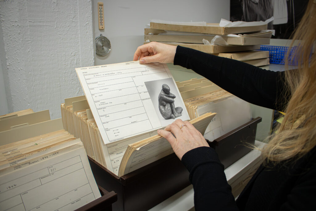 Närbild av ett verkspapper i ungefär A4-storlek som Mariaana Korpela studerar. På verkspappret finns ett svartvitt fotografi av samma skulptur i röd lera som i början av artikeln vid rengöringen. Dessutom står verkets uppgifter angivna.