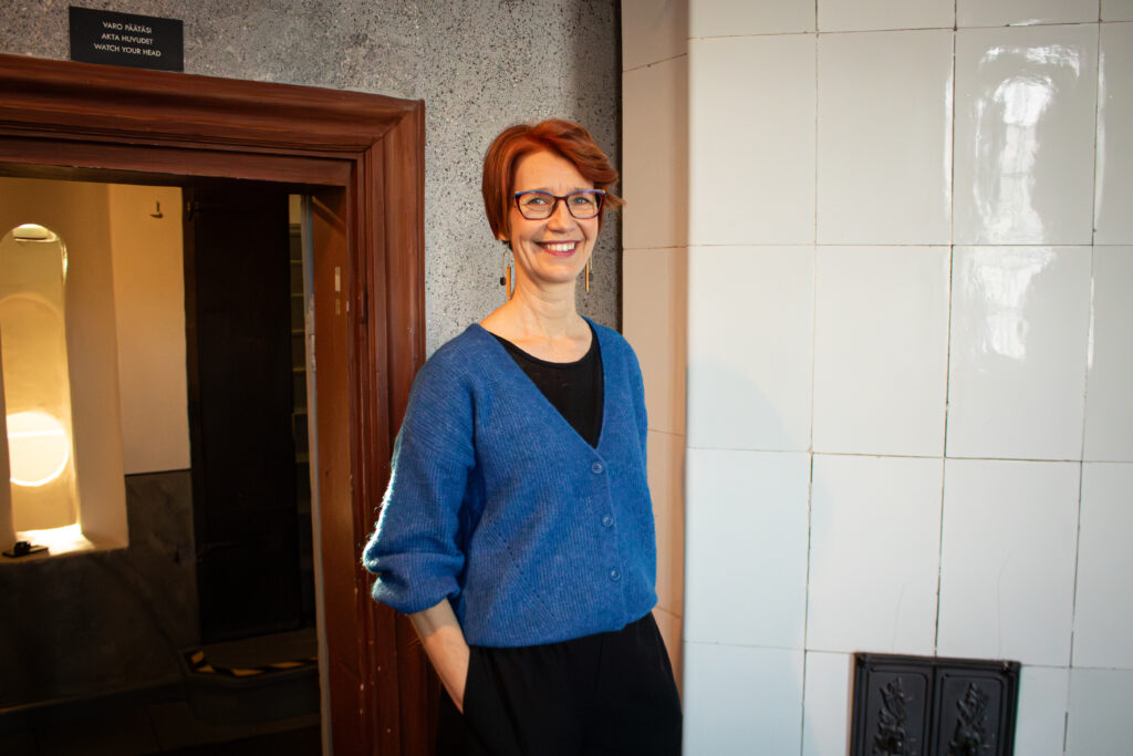 Iina Wahlström står i dörröppningen, ler mot kameran. I bakgrunden syns en vit kakelugn samt en korridor som öppnar sig från dörröppningen.