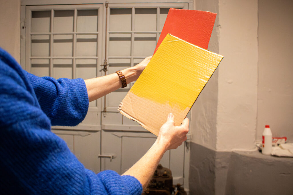 Iina Wahlström pitää kädessään kahta pahvilevyä. Etualalla oleva pahvi on maalattu keltaiseksi ja taka-alalla oleva punaiseksi.