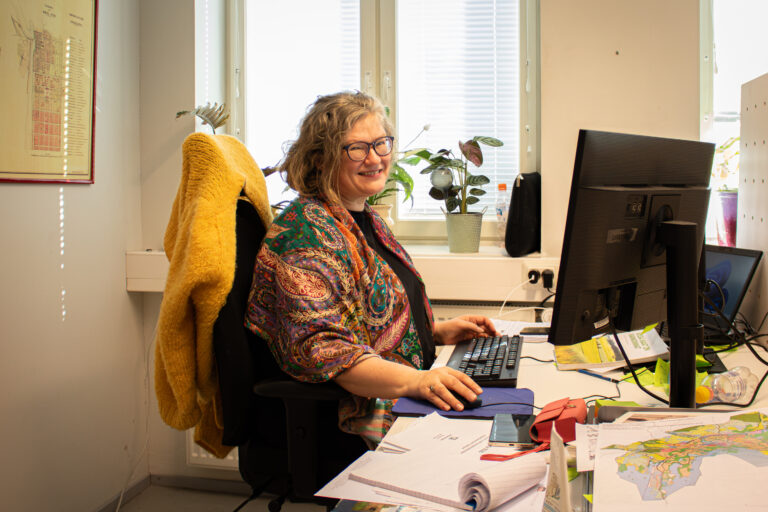 Hannele Lehtinen sitter vid datorn på kontoret och ler mot kameran.