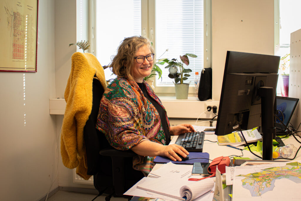 Hannele Lehtinen istuu toimistossa tietokoneen äärellä ja hymyilee kohti kameraa.