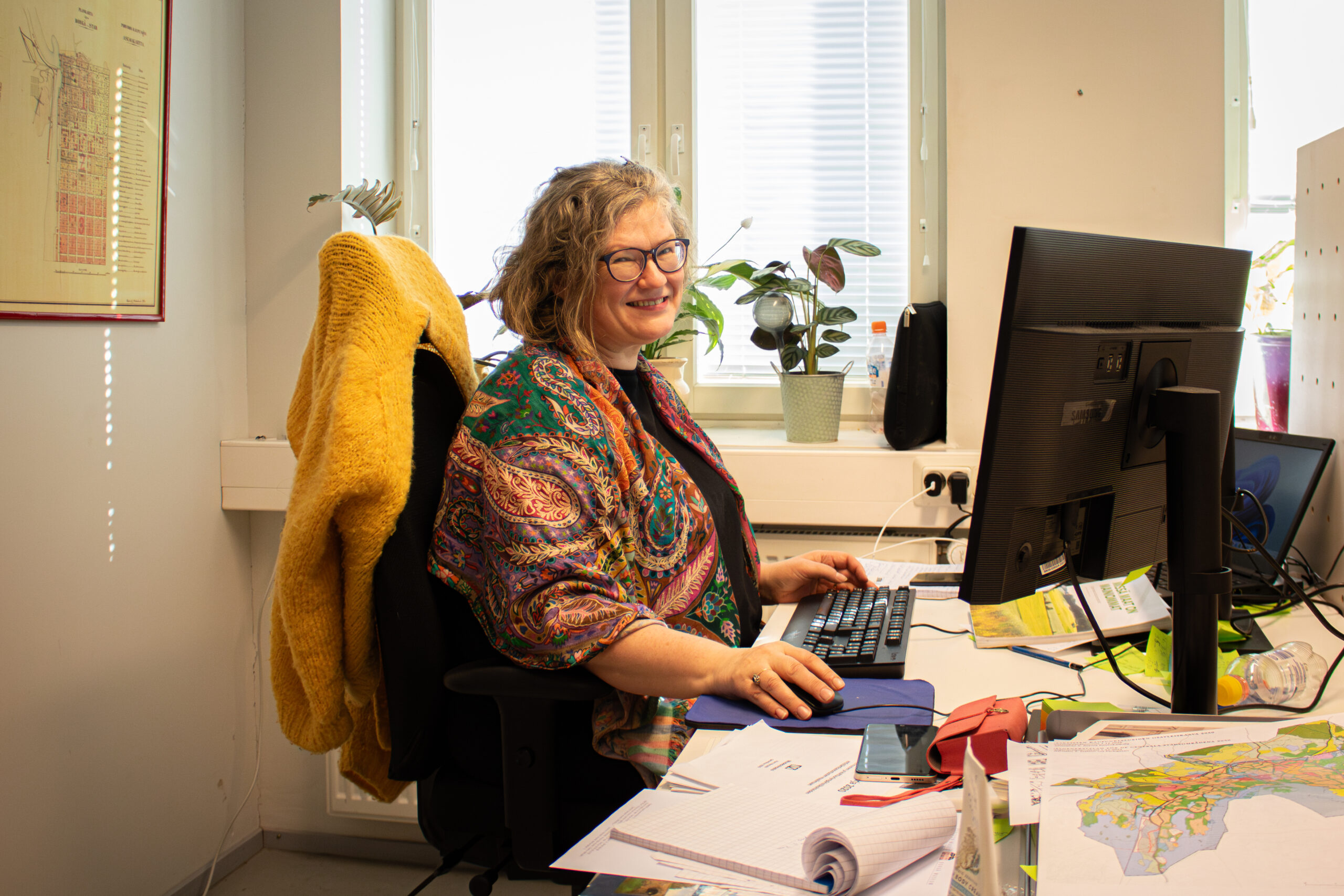 Hannele Lehtinen istuu toimistossa tietokoneen äärellä ja hymyilee kohti kameraa.