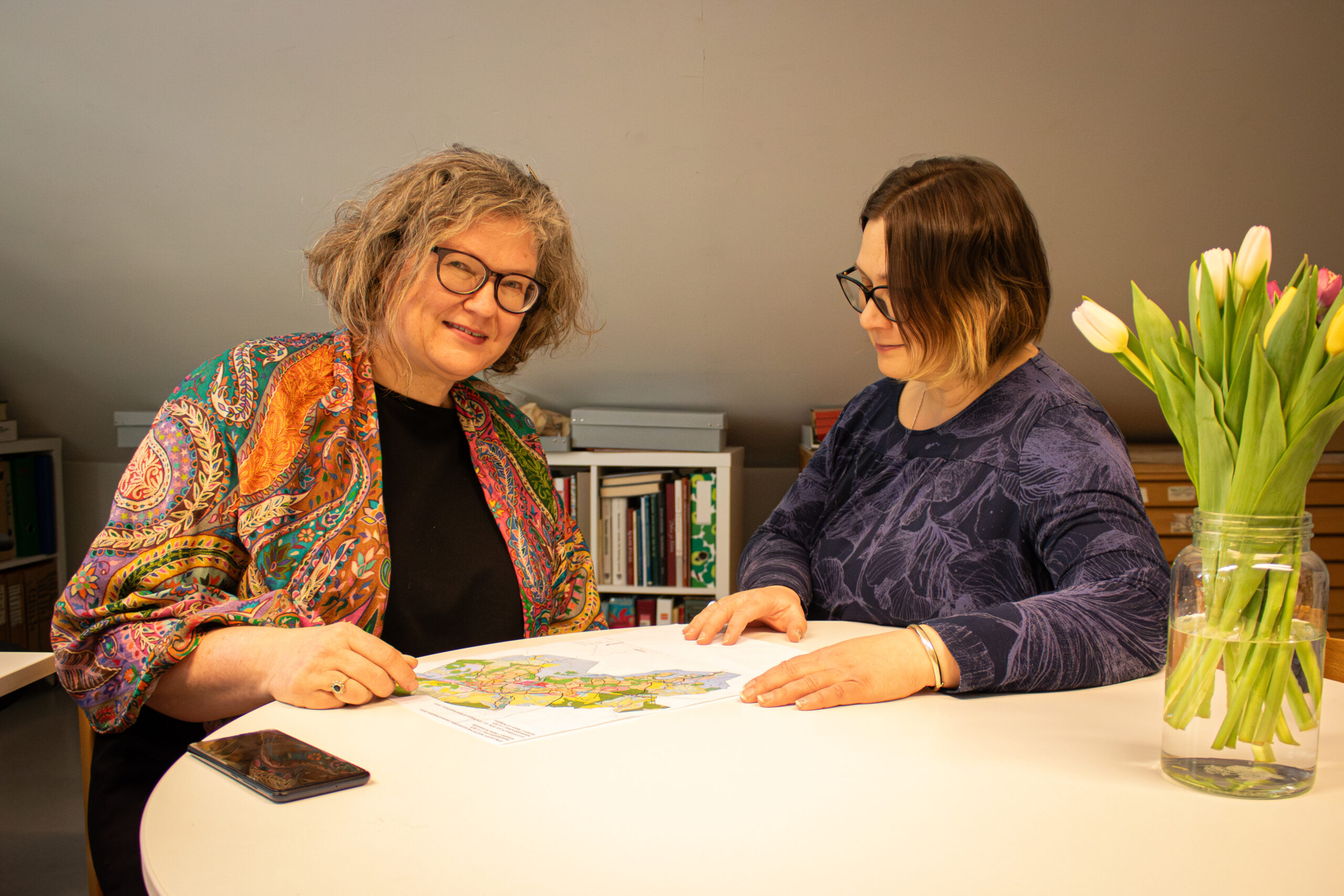 Hannele Lehtinen och arkeologen Riikka Mustonen sitter vid ett runt vitt bord och studerar delgeneralplanen tillsammans. Lehtinen sitter till höger och ser mot kameran. Mustonens blick är fäst vid planen. På bordet står gula och röda tulpaner i en glasburk.