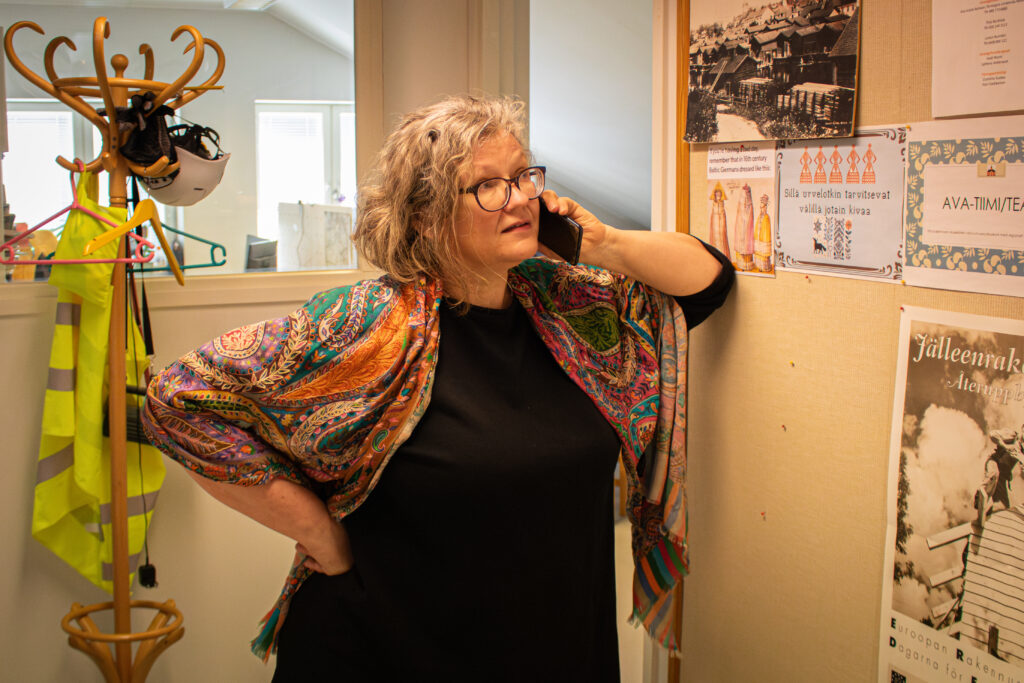 Hannele Lehtinen talar i telefon i kontorskorridoren och lutar sin vänstra armbåge mot en anslagstavla som är fäst vid väggen. Lehtinen tittar upp mot anslagstavlan.
