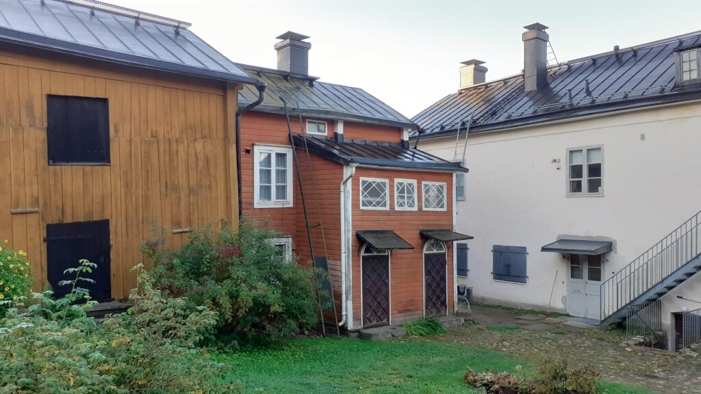 Kuvassa on kolme rakennusta. Oikealla puolella näkyy keltamullanvärinen puurakennus, keskellä punertavanoranssi puurakennus eli Wikmanin talo ja vasemmalla puolella kuvaa näkyy valkoinen kivitalo eli Holmin talo. Kuva on otettu Holmin talon pihapiiristä. Kuvassa näkyy lisäksi vihreää ruohoa ja muutama pensas.