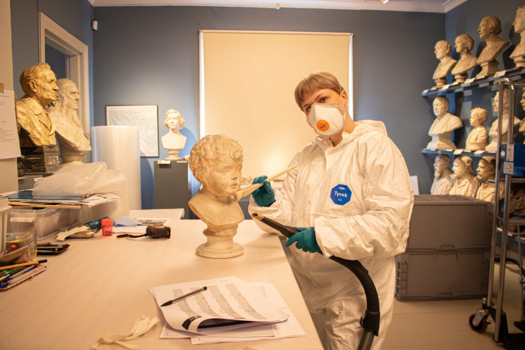 Nea Satta puhdistaa valkoista kipsiveistosta, joka esittää lapsen päätä. Satalla on päällään suojavarusteet ja yhdessä kädessä ihmurin suurit ja toisessa sivellin. Hän katsoo suoraan kuvaa kohti, pää hieman kalleellaan.