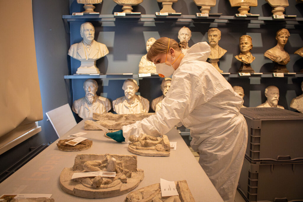 Nea Satta kontrollerar inventarienumren på skulpturerna som ligger på bordet, skrivna på små papperslappar. Hon bär vita skyddsoveraller, skyddsmask och blå handskar. På hyllan bakom Satta syns vita skulpturer mot en blå vägg.