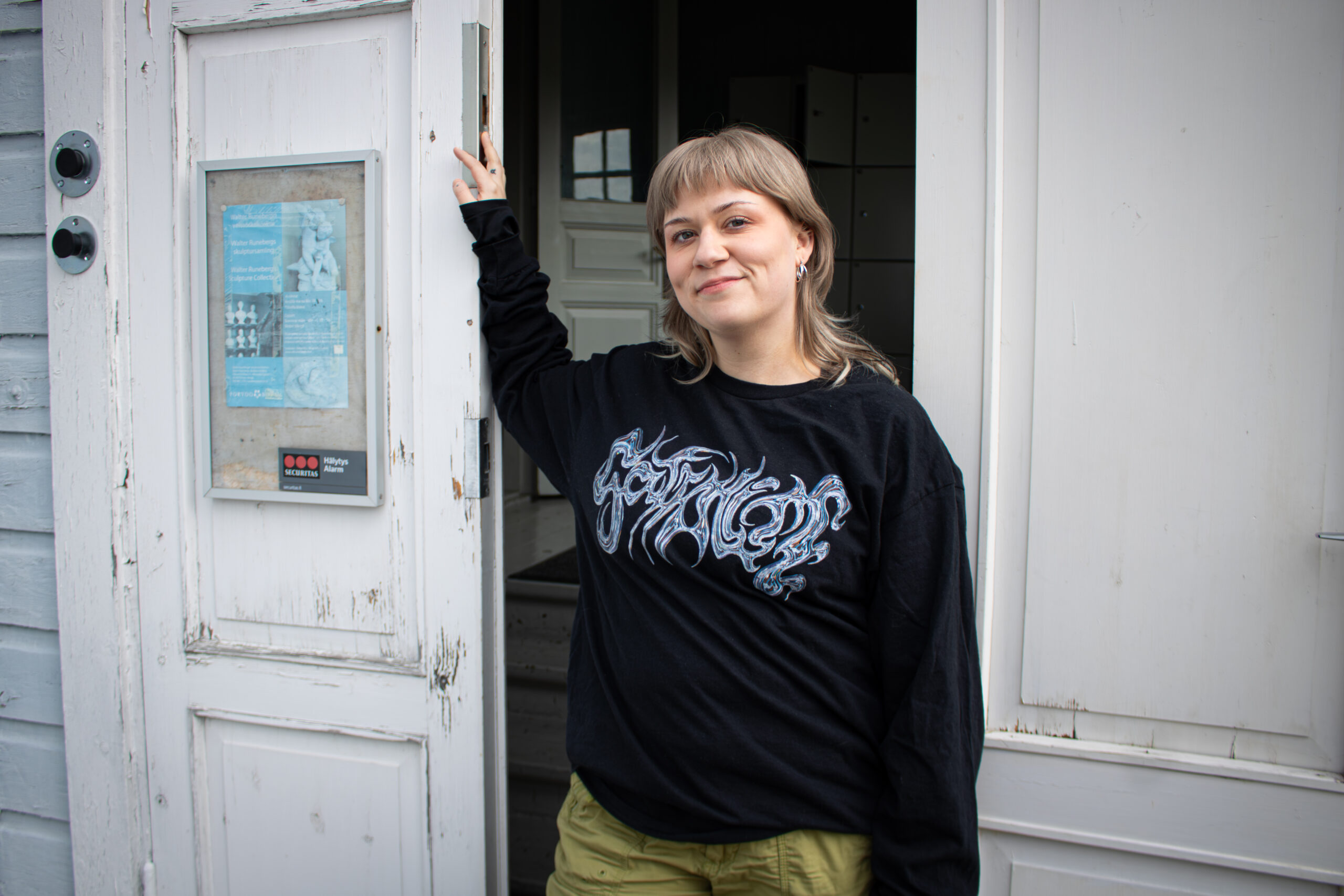 Suoraan kameran kuvaan katsova Nea Satta seisoo vanhan sinisen värisen hirsitalon etuovella ja nojaa oikealla kädellä valkoista puuovea vasten.