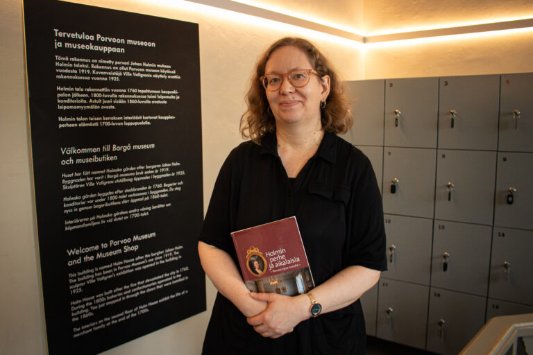 Porvoon museon näyttelyvalvoja ja opas, taiteilija Sanna Johansson seisoo ja katsoo hymyillen. Hänellä on kädessään punainen kirja. Taustalla oikealla on harmaita säilytyslokeroita ja vasemmalla teksi mustalla pohjalla ja valkoisella tekstillä, missä asiakkaat toivotetaan tervetulleeksi Porvoon museoon.