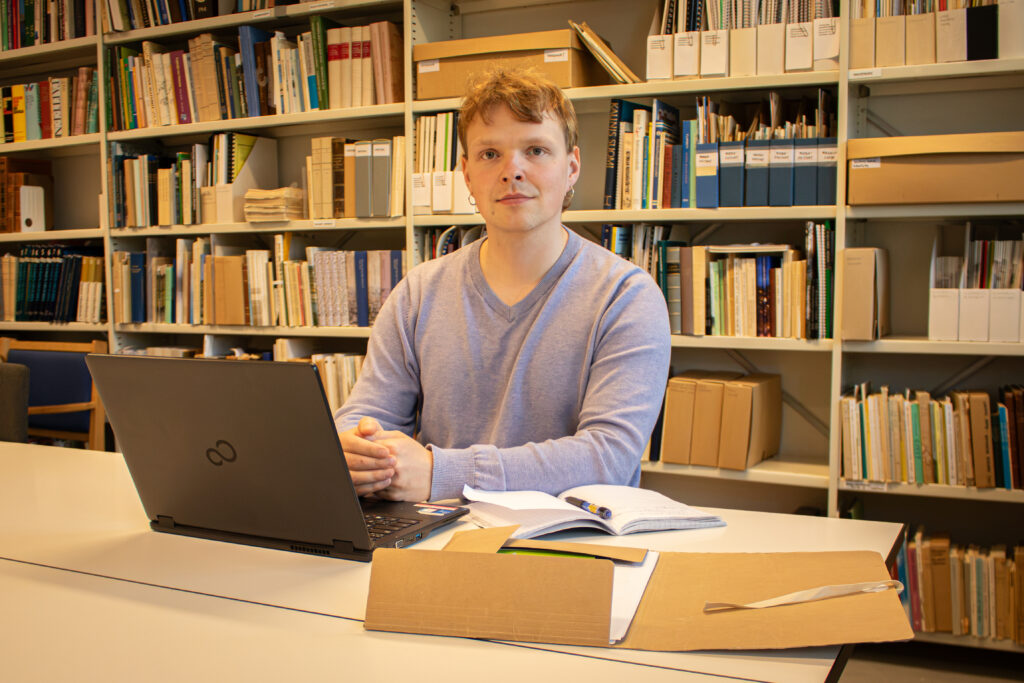 Olli Järveläinen istuu kohtisuorassa katsojaan päin valkoisen pöydän äärellä, jossa on musta kannettava tietokone, ruskea kansio. jossa on papereita, sekä muistiinpanovälineet. Taustalla iso ja valkoinen metallihylly, jossa paljon erilaisia kirjoja.