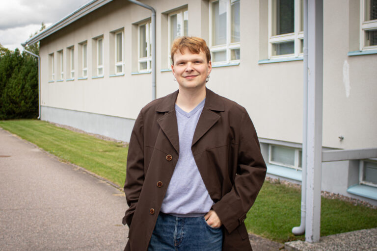 Byggnadsforskare Olli Järveläinen står utomhus i en brun jacka, en lila skjorta och mörkblå jeans och ler mot kameran. I bakgrunden syns Borgå museums vita stenbyggnad, som inrymmer museets kontor. På bilden syns också en grön gräsmatta och buskar, asfalt samt en molnig himmel.