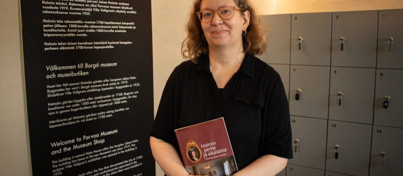 NYTTEL~2 Borgå museums utställningsvakt och guide, konstnären Sanna Johansson, står och ser leende mot kameran. I handen håller hon en röd bok. I bakgrunden syns till höger grå förvaringsskåp och till vänster en svart skylt med vit text som hälsar besökarna välkomna till Borgå museum.